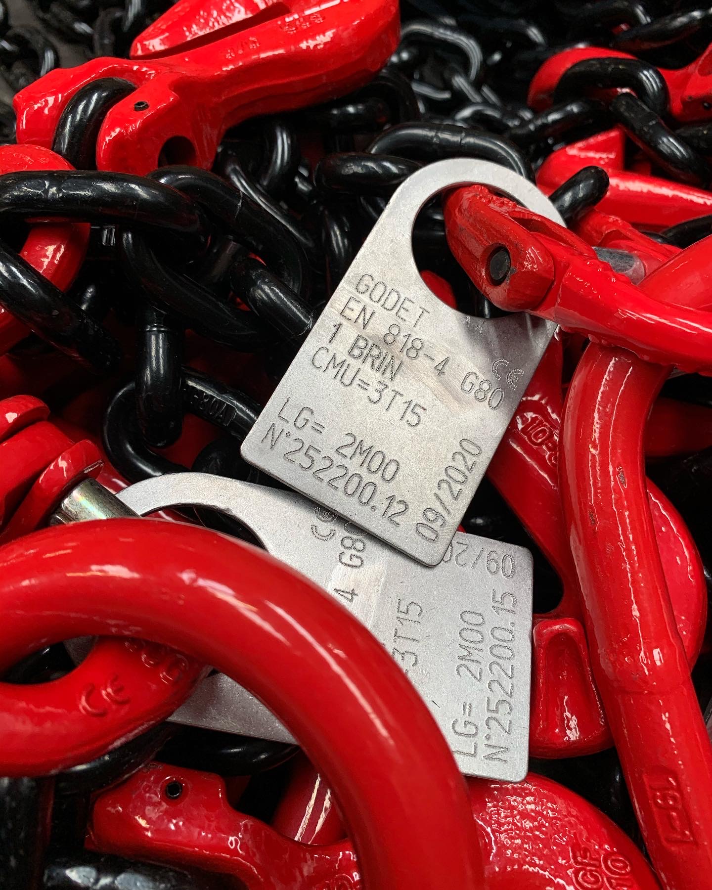 Éléments de chaînes de levage en acier noir et crochets rouges, avec plaques d’identification métalliques gravées indiquant les normes et capacités de charge.